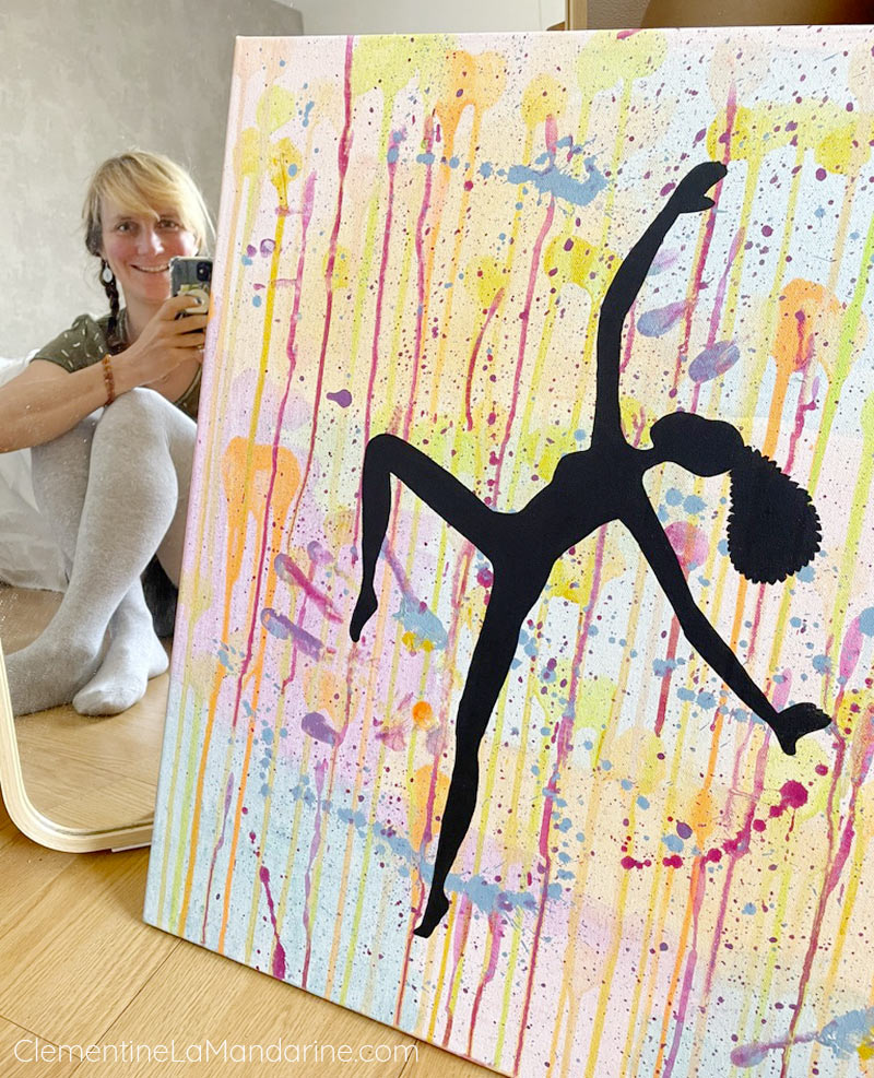 Une personne souriante derrière un tableau coloré, avec une silhouette de danseuse en noir.